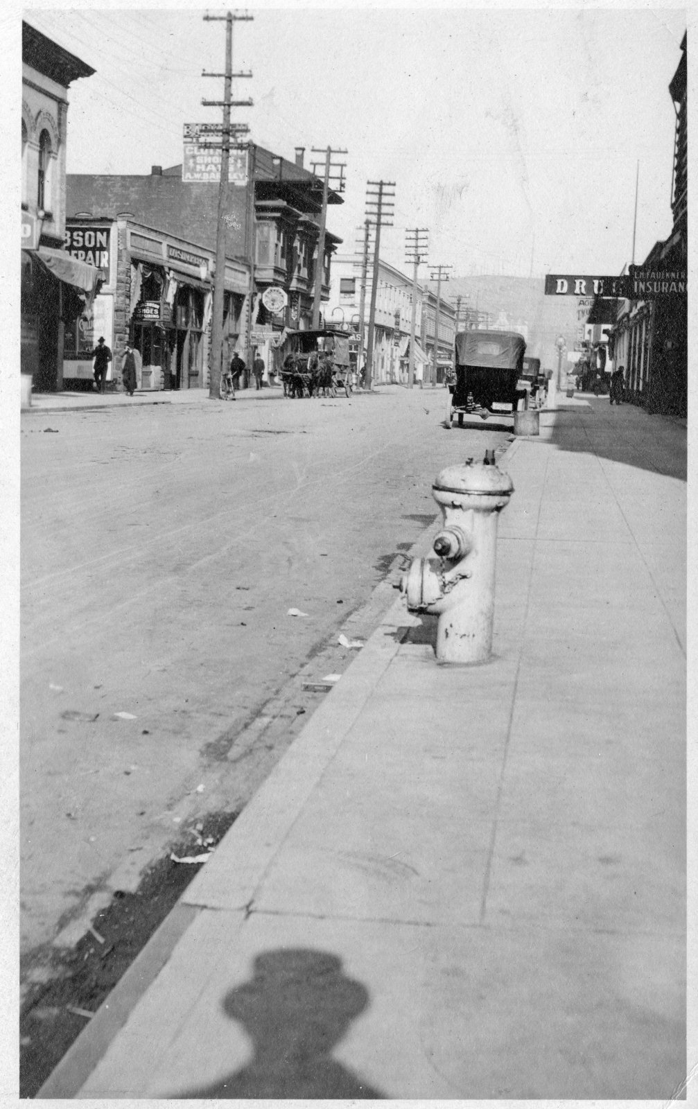 A black and white street scene with horse-drawn carriages and early automobiles, likely from the early 20th century.