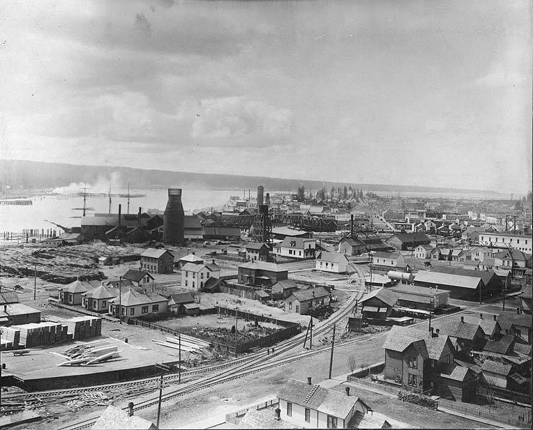 A black and white photograph of a bustling industrial and residential area, likely in the late 19th or early 20th century, with visible steamships and industrial structures.
