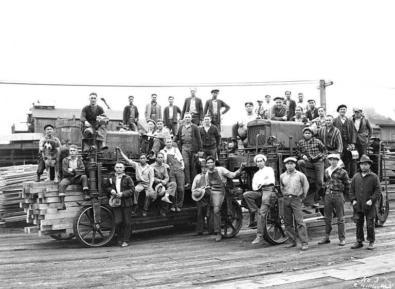 A group of workers posing with a steam-powered logging machine in a lumberyard.